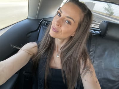 A woman with long, brown hair is taking a selfie from the backseat of a car. She is wearing a black tank top and a necklace with a dragonfly pendant. Her left arm is visible, showing a tattoo. The interior of the car is dark leather. Through the window, a blurred road and trees are visible, indicating movement. The scene suggests the woman is traveling, likely on a sunny day.