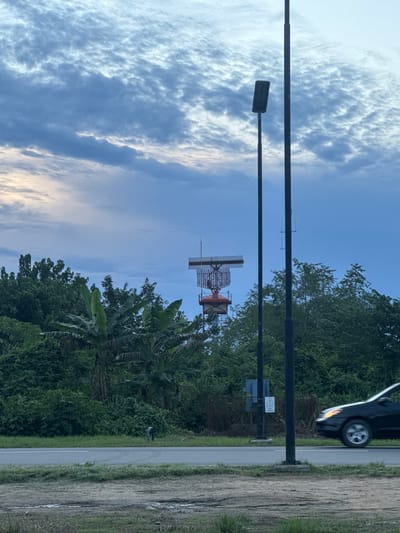 The image depicts a rural scene with a blue sky covered in scattered clouds. A black car is partially visible on the right side of the road. Alongside the road, there's lush green vegetation interspersed with tall trees and banana trees. In the distance, a red and white radar tower is seen above the treeline. Two black lampposts and electrical poles are visible in the foreground between the sky and the trees. The image captures a moment in Omoecheigbo, Nigeria, showcasing a blend of natural elements and infrastructure.