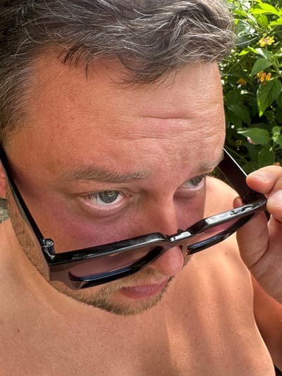 This media file is a close-up, rotated photograph of a person's face. The subject is a fair-skinned individual with short, graying brown hair. They are wearing dark-rimmed sunglasses, which are being held up by a hand. The person's right eye is visible, looking directly at the camera. The skin shows some texture and is slightly flushed. There is stubble on the chin and jawline. The background is blurred and appears to be foliage with some small yellow flowers, suggesting an outdoor setting in daylight. The lighting indicates it is likely daytime, possibly sunny. The overall mood is casual and candid. There is no visible text in the image.