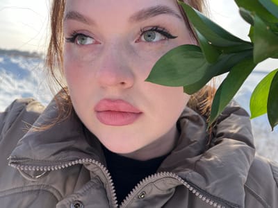 A close-up photograph depicts a young adult female subject outdoors in Pavlovka, Russia, against a snowy landscape under bright sunlight. The subject has reddish-brown hair, fair skin with visible freckles and flushed cheeks, light green eyes, and full lips with light pink lipstick. Her left eye (viewer's right) is fully visible, featuring black winged eyeliner and prominent eyelashes, while her right eye (viewer's left) is partially obscured by a cluster of vibrant green leaves from an unidentified plant branch held in the foreground. She is observed looking upwards and to her left (viewer's right). The subject wears a light brown or beige puffer jacket over a black garment, with a zipper visible on the jacket's front. The background consists of an expanse of snow-covered ground extending to a distant horizon under a clear, bright sky, indicative of a sunny winter day.
