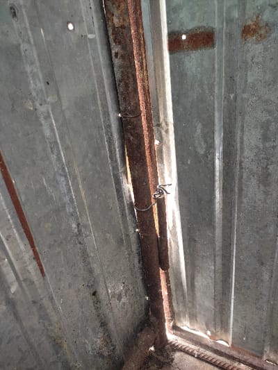 The image shows a close-up of a rusty metal hinge attaching a corrugated metal door to a metal frame. The hinge is secured with a metal wire. The image is likely taken in a rural area or a neighborhood in Kegeti, Kyrgyzstan, based on the coordinates provided. The scene suggests a simple, functional, and potentially aging structure in a less-developed area.