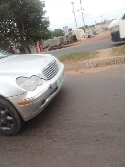 A silver Mercedes-Benz is parked on the right side of a paved road in the city of Abuja, Federal Capital Territory, Nigeria. The car is facing towards the left side of the image. The road is relatively empty and there are power lines overhead. The side of the road near the car has a sidewalk with green grass growing on it. There is a white van in the background, partially visible, and a man is walking on the road in the distance. There is a sign that says "350 DESIGN" written on the curb near the car. The weather appears to be cloudy.