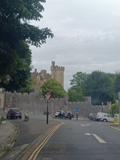 The image shows a view from a road with a castle in the background. The castle is made of stone and has turrets. A stone wall surrounds the castle grounds. Trees and greenery are visible throughout the scene. There are several cars parked on the road and a give way sign is visible. The sky is overcast. The location is identified as Arundel, United Kingdom.