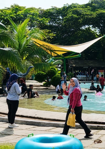 The image depicts a sunny day at a water park, likely located in Pantai Cermin Kiri, Indonesia, given the presence of lush tropical foliage and a relaxed, communal atmosphere. In the foreground, a woman in a pink long-sleeved shirt and black pants, wearing a light blue and purple hijab, waves at the camera. She carries a yellow shopping bag in her right hand. Behind her, a large blue inflatable ring rests on the paved ground.
The midground features a wading pool with clear, shallow water. Several individuals are visible in the pool, including children splashing. Some adults are seated on the pool's edge or nearby. To the left of the pool, another woman in a striped long-sleeved shirt and black pants, with a blue hijab, stands facing away from the camera. In the background, palm trees and other dense green foliage create a tropical environment. A distinctive yellow and white sunshade structure is visible above the pool area, supported by green poles. Beyond this, a wooden building with a dark roof can be seen, suggesting amenities such as changing rooms or a cafe. The overall scene conveys a sense of leisure and family recreation.