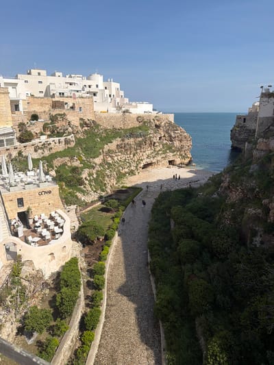 The image depicts a coastal scene in Polignano a Mare, Italy. In the foreground, there's a restaurant terrace with white tables and chairs. Adjacent to the terrace, a stone wall separates it from a pathway made of small stone pieces. The pathway curves along the edge of a steep cliff covered in greenery. Below the cliff, a small beach is visible, populated with a few individuals. The sea stretches out beyond the beach, meeting the horizon under a clear blue sky. On the left side of the image, there are clusters of white buildings clinging to the cliffside. The overall perspective is from an elevated viewpoint, likely a building or higher ground overlooking the beach and sea.