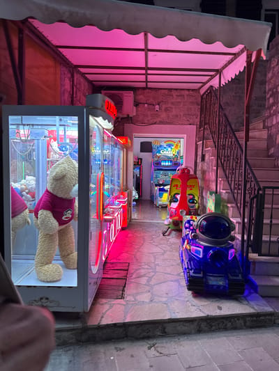 The scene is an outdoor arcade in Kaluđerac, Montenegro. The arcade is located on a stone pathway, which leads up to a set of stairs with wrought iron railings. Along the pathway, there are several arcade machines. The closest machine is a claw crane game filled with stuffed animals. Behind it are additional arcade games, including driving simulators and a "Crazy Taxi" game. An awning, lit with pink lights, covers the left side of the scene. An air conditioning unit is visible on the wall behind the awning. The arcade is surrounded by stone walls. The image also captures a hand holding a device in the bottom right corner.