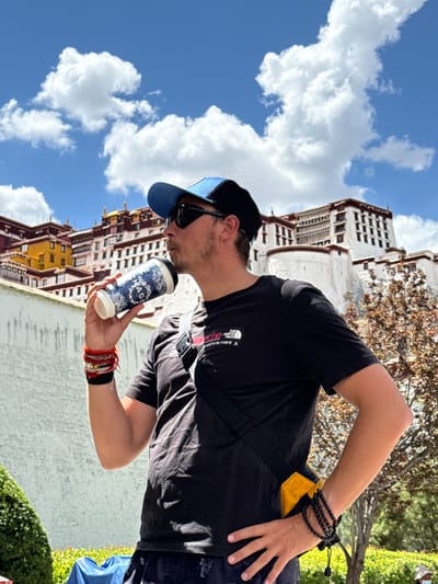 A person is captured in a sunlit outdoor scene, holding a white and blue disposable coffee cup to their lips, appearing to take a sip. The individual is positioned on the right side of the frame, seen from the waist up, and is wearing a black short-sleeved t-shirt, a blue baseball cap, and sunglasses. Their right wrist is adorned with multiple red and orange string bracelets and a wider black one, while their left wrist features several black beaded bracelets. A small yellow pouch with a black strap is visible near their left hip.
In the background, a majestic, multi-story building, characteristic of the Potala Palace, rises prominently. It features distinct white walls, red and yellow sections, and numerous windows, suggesting an ancient and grand architectural style. Lush green foliage and trees are visible around the building and in the foreground. The sky overhead is a vibrant blue, dotted with fluffy white clouds, indicating clear weather.
The activity appears to be a casual moment of refreshment or relaxation, with the person enjoying a beverage amidst significant historical surroundings. The time of day is clearly daytime, with bright sunshine.
Visible text includes a logo on the person's black t-shirt which reads "THE NORTH FACE" along with "NEVER STOP EXPLORING" and coordinates "N°39.87106°E" and "N°116.39758°E". On the coffee cup, a circular logo displays the word "AGE" centrally, surrounded by a blue and white decorative pattern. Given the architectural style and the provided location context, the scene is undoubtedly set in Chengguan District, China, with the Potala Palace as a prominent backdrop.