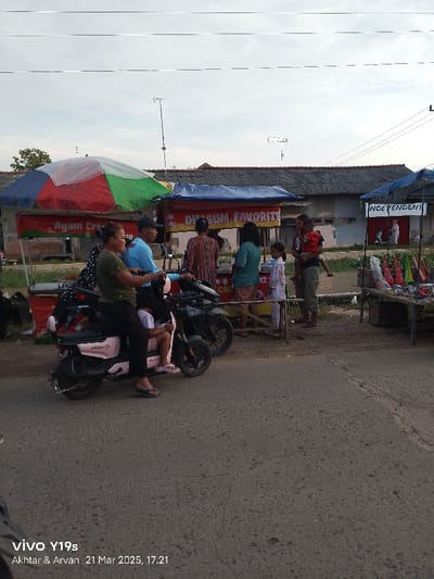The image depicts a scene on a street in the city of Kecamatan Kroya, Indonesia, located at latitude -6.422 and longitude 107.996. The street is paved and appears to be relatively narrow. The image is taken from the perspective of someone standing on the street, looking towards the right. Several people are present in the scene, including a group of individuals standing in front of a food stall, and a woman on a pink electric scooter. There are also various objects, including a scooter, a food stall, a small child holding a purse, and a nearby street vendor selling brightly colored objects. The food stall has a blue awning and a sign that says "Dimsum Favorite". The woman on the scooter is wearing a blue shirt and black pants, and is holding the handlebars. She appears to be in the process of driving the scooter forward. The small child in the scene is standing next to the scooter, and is wearing a white shirt and blue pants. The street vendor is standing behind a small table and has a variety of items for sale. The environment is characterized by the presence of buildings, trees, power lines, and a dusty, open road. The image is shot under overcast skies.
