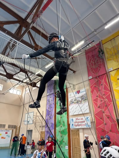 A person wearing a helmet, dark athletic clothing, and gloves is suspended mid-air, attached to a rope course within an indoor structure. The person is wearing a harness, and the cables are attached to it. The background reveals a high-ceilinged interior with a climbing wall decorated with various colors (yellow, red, green, and purple). There are posters on the climbing wall with writing in Russian. There is a basketball hoop mounted on a wall. Several other people stand on the ground, watching. Based on the location of the signs, the image was taken in Orel, Russia.