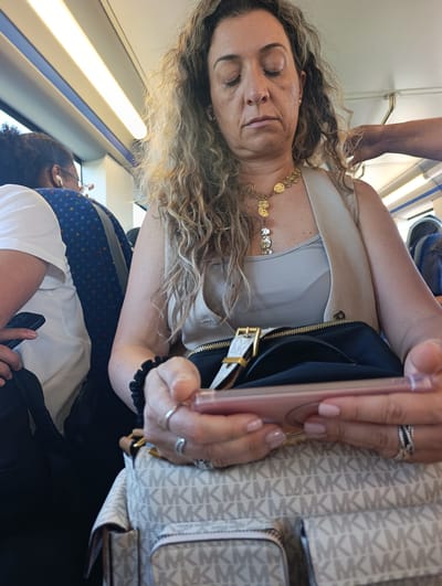 A woman with curly brown hair, wearing a light-colored sleeveless vest over a matching tank top and a gold necklace, sits on a train. She is holding a pink smartphone in her hands. Her eyes are closed, and she appears to be resting or listening to something through wireless earbuds. In front of her is a black bag with gold hardware and a white strap with a repeating "MK" logo, possibly a Michael Kors bag. She is wearing rings on her fingers. To her left, another passenger wearing a white t-shirt and glasses is visible, also with earbuds. The train interior features blue and white striped seats and overhead fluorescent lighting. The image is a close-up shot, focusing on the woman and her immediate surroundings. The scene is consistent with being aboard a public transportation vehicle, likely a train, during daylight hours. The location is not discernable from the image itself, but given the context of the prompt, it is stated to be Palmela, Portugal.