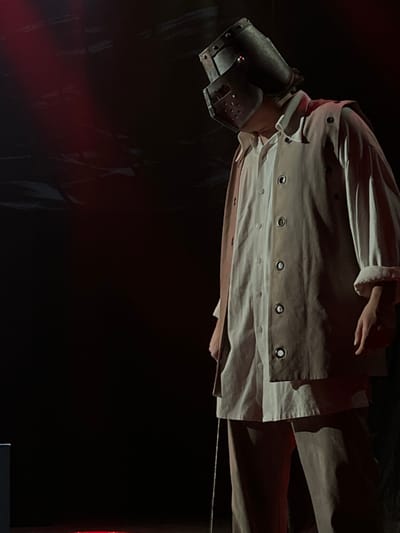 A single individual is observed standing on a dark stage in Novoshakhtinsk, Russia. The person, viewed from the mid-thigh upwards, is illuminated by directed red stage lighting from the upper left. They are wearing a long-sleeved, light-colored shirt or tunic featuring multiple large, dark circular grommets arranged vertically down the front, over matching light-colored trousers. A dark, riveted metal helmet, resembling a medieval great helm with narrow eye slits and perforated breathing holes, encases the head. A distinct small red light source is visible within the left eye slit of the helmet. The subject's head is bowed downwards. A slender, reflective metallic rod is positioned vertically alongside the individual's lower right side, extending out of the frame downwards. The background is entirely black, with subtle, indistinct dark textures visible in the upper left quadrant. No other persons or specific objects are identifiable in the immediate environment.