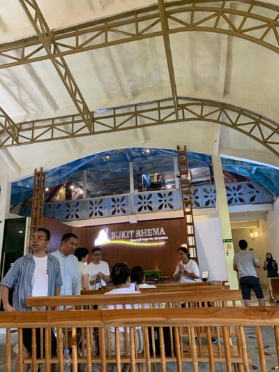 The image depicts a scene inside a building with a large, open, wood-framed ceiling structure, and a balcony on the second level. A wooden sign reads "BUKIT RHEMA" in large lettering. The sign is located on the lower level wall next to a small doorway. The building is a house of worship, with a mix of people standing and sitting on wooden benches. There are at least six people present: one man in a striped shirt and a woman with her hands clasped in prayer are standing near the entrance, one man in a grey shirt is standing near a woman in a white top, and two people seated on the bench are partially obscured. The balcony above the entrance is decorated with blue walls featuring a depiction of clouds, and a pattern of white stylized flowers. The balcony is accessible by an unlatched wooden ladder. The location appears to be a house of worship in the Kecamatan Borobudur, Indonesia, with coordinates -7.606 latitude and 110.181 longitude.