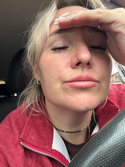 The image is a close-up, waist-up, selfie-style photograph of a young woman inside a vehicle. The woman has fair skin, blonde hair, and is wearing makeup, including false eyelashes. She is wearing a red zippered jacket over a brown shirt, and a necklace with black and gold beads. Her left hand is pressed against her forehead. The interior of the car is visible, including a steering wheel, car seat, and a part of the dashboard. The background is blurred and appears to be outdoors, possibly in a residential area with buildings and trees. The exact location in Chastyye, Russia, cannot be determined from the visual information provided.