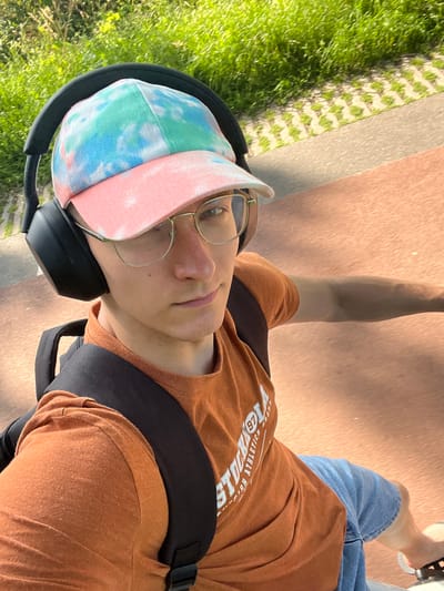 The image is a selfie of a young man with glasses and headphones, wearing a tie-dye baseball cap, an orange t-shirt, and denim shorts. He has a backpack on. He appears to be outside, possibly in a park or along a paved path, with grass and vegetation visible in the background. The ground in the foreground is a reddish-brown color, suggesting it could be a bike path or walkway. The lighting indicates it is daytime. The specific location within the city of Enschede, Netherlands cannot be determined from the visual information provided in the image.