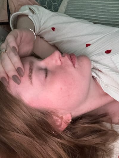 The image displays an individual, likely a young adult female, lying on a surface consistent with a bed or sofa, within an indoor environment located in Rosmalen, Netherlands. The subject is positioned in profile, facing left, with her eyes closed and lips slightly parted, indicating a state of rest or sleep. Her complexion presents with a reddish hue. Long, light brown hair is visible, spread across the surface around her head. Her left hand is placed on her forehead; the fingers exhibit long, coffin-shaped nails painted a matte taupe color. A ring with a clear stone is visible on the ring finger of the left hand, and a silver-colored bracelet with multiple small reflective elements encircles her left wrist. She is dressed in a white, long-sleeved garment, possibly a shirt, which features scattered small red heart-shaped motifs. The background is out of focus, revealing soft furnishings, including pillows, one with a geometric pattern and another with horizontal stripes, in muted shades of blue-green and grey.