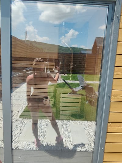 A person is captured in a reflection on a glass window. The reflection shows the person from the waist down, standing on a path paved with white pebbles. They are wearing pink clogs. In the background, through the glass, a wooden structure with horizontal planks is visible, along with a grassy area. Sunlight illuminates the scene, casting shadows on the ground. The reflection also shows elements of the sky, with white clouds against a blue background, and a distant hill covered with greenery and some wooden structures. A small wooden bucket and ladle are visible near the person in the reflection. The image appears to be taken outdoors, possibly in a backyard or garden area. The exact location in Zheleznovodsk, Russia cannot be determined from the visual information provided.