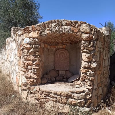 The image depicts an outdoor shrine constructed from rough-hewn stones, likely located in the vicinity of Terlizzi, Italy, given the context of the photograph. The shrine is built into a curved wall, with a recessed niche at its center. Within the niche, a carved stone bas-relief depicts a figure with a halo and raised hands, resembling a religious icon. The shrine is weathered, with some stones displaced and the surrounding area overgrown with dry grass and some sparse vegetation. A small, circular ceramic tile with an image of flowers is affixed to the stone wall to the left of the niche. The sky above is clear and blue, indicating a sunny day. The timestamp "26.06.2026 10:42" is visible in the lower right corner of the image, suggesting it was taken on June 26, 2026. There are no people or animals visible in the frame.