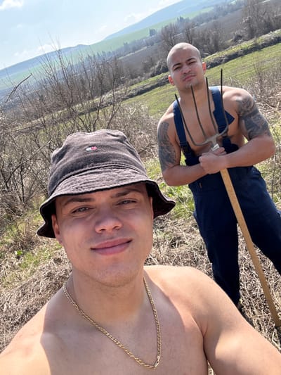 This image captures two males outdoors in Katselovo, Bulgaria, under bright, clear conditions. In the foreground, a male with a light complexion and short hair wears a dark bucket hat with a small emblem and a gold-colored chain necklace. He is shirtless and looks directly at the camera with a slight smile. Behind him, positioned slightly higher and to the right, stands another male with a shaved head and light complexion. This individual is shirtless, revealing extensive dark tattoos on both arms, and wears dark blue overalls. He holds a three-tined pitchfork with a wooden handle, pointing upwards. His expression is neutral as he looks towards the camera. The immediate environment consists of dry, light brown grass and sparse, leafless bushes. In the background, rolling green hills extend under a clear blue sky.