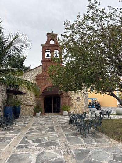 This image captures an outdoor courtyard scene, likely in a historic or cultural setting. Dominating the frame is an old brick building with a bell tower, featuring arched openings and several bells. Adjacent to the building is a stone-paved pathway laid with irregular grey flagstones. To the left of the building, a large tree with dense green foliage shades the area. In the background, a yellow building is visible.
Several dark metal chairs are neatly arranged on the stone patio. A small blue golf cart or utility vehicle is parked near the yellow building. In the lower foreground, a palm frond is prominent, suggesting a tropical or subtropical location. There are also large blue cylindrical planters and some greenery. The sky is overcast, indicating a cloudy or possibly rainy day.
A sign on the stone wall of the building reads "CASA DON LUIS", "CAP CANA", and "BY MARIOTT BOUTIQUE". This text clearly indicates the location as Cap Cana, a resort area in the Dominican Republic, and identifies the building as Casa Don Luis, part of a Marriott Boutique property. The overall mood is serene and perhaps a bit somber due to the overcast weather. The architecture suggests a blend of colonial and tropical influences. No people are visible in the image.