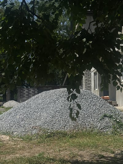 The image shows a large pile of grey gravel in front of a building under construction in Yablunivka, Ukraine. Overhanging dark green tree branches partially obscure the view of the building and the sky. The building appears to have a section with stone-like siding and smooth grey walls with white-framed windows. To the left of the gravel pile, there is a stone wall with a brick pattern. The ground in the foreground consists of dry dirt and patches of green grass. A small toy dump truck is visible on the steps leading to the building. The scene appears to be a construction site with building materials and partially completed construction.