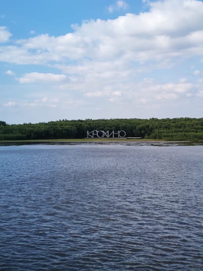 The image shows a body of water in front of a densely wooded shoreline. A large, white, three-dimensional inscription spelling out "КРОХИНО" (Krokhino) is placed on the shore, centered in the frame and partially submerged. The inscription's letters are blocky and stylized. The water is dark blue and appears somewhat choppy with small ripples. The shoreline is lined with dark green trees that form a solid wall of foliage. Above the trees, a bright blue sky is visible, populated by scattered white, fluffy clouds. The overall scene suggests a natural landscape with a prominent man-made landmark.