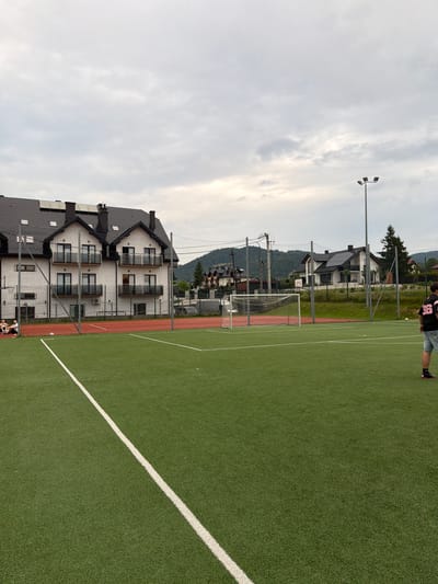 The image depicts an outdoor artificial turf soccer field in Mordarka, Poland, under an overcast sky. A white line marking the edge of the field runs diagonally from the bottom left to the top center of the frame. In the background, there is a multi-story building with a dark-tiled roof and balconies. To the right of this building, a soccer goal with a net is visible. Further in the background, more buildings and a wooded hillside are present. On the left side of the field, two individuals are seated near a fence. On the right side of the field, a person wearing a black jersey with the number "36" in pink is standing. A soccer ball is visible on the grass near the goal.