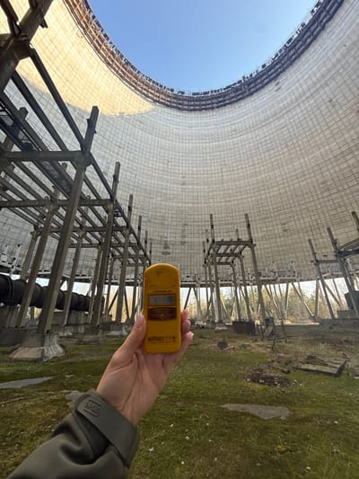 The image depicts the interior of one of the cooling towers within the Chernobyl Nuclear Power Plant Exclusion Zone in Pryp'yat', Ukraine. A person is holding a yellow radiation detector, a "TERRA-P" dosimeter manufactured by "ECOTEST," pointing upwards towards the tower's interior. The tower is constructed of concrete blocks forming a curved, open structure with visible metal support beams and scaffolding. The sky is visible through the top of the tower. The ground surrounding the base of the structure is overgrown with grass and vegetation, including scattered debris. The person's hand and a portion of their jacket are visible, indicating they are wearing outdoor clothing.