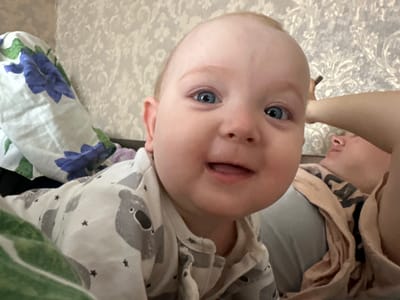 A close-up, selfie-style shot captures a fair-skinned baby with blue eyes and light blonde hair smiling. The baby is wearing a pajama set featuring a koala bear pattern and small stars. To the left of the baby, a white cloth with large blue flowers and green leaves is visible. In the background, a textured, light-colored wallpaper adorns the wall. To the right, an adult's arm is extended, and a person's head is resting on their chest, partially obscured. The scene appears to be indoors, possibly in a home setting.