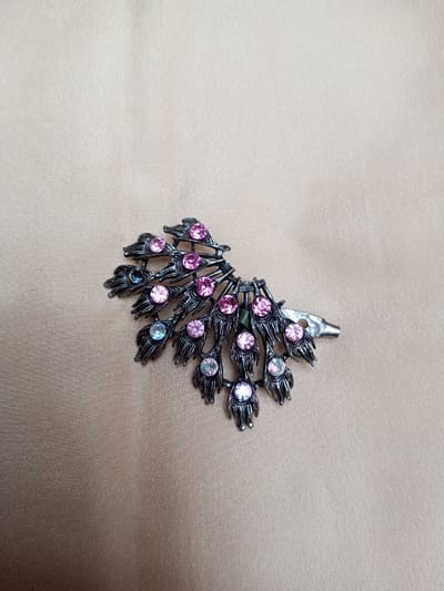 A dark metallic hair clip, stylized as a peacock feather, is positioned diagonally on a light beige fabric surface. The clip, oriented with its fanned portion extending to the left and the clasp mechanism to the right, features approximately fifteen round pink rhinestones and three round light blue rhinestones embedded within its textured, segmented design. The metal exhibits a dark, possibly gunmetal or oxidized finish. The background fabric displays subtle folds and a slight sheen. The scene is located in Qarabucaq qəsəbə, Azerbaijan.