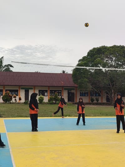 The image depicts a volleyball game in progress, likely on a school's outdoor court in Blangriek, Indonesia. Four young female athletes, dressed in black hijabs and orange sportswear, are visible on the court. One player is in mid-action, about to hit the ball, while the others are positioned around the court, anticipating the play. The court itself is brightly colored, with distinct yellow and blue sections. Beyond the court, a volleyball net stretches across the frame, separating the players. In the background, a building with a reddish-brown roof and a row of windows is visible, typical of Indonesian school architecture. Large trees with lush green foliage stand behind the building, and a cloudy sky completes the scene.