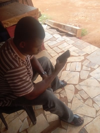 A man is sitting on a black plastic chair outdoors, looking at a dark-colored smartphone he is holding in his right hand. He is wearing a brown and white striped polo shirt, dark gray pants, and black slide sandals. He has a watch on his left wrist. The ground is covered with irregularly shaped beige and white stone tiles. Behind the man is a table with items covered with a brown cloth. In the background, there is a raised border of stone tiles separating the tiled area from a reddish-brown dirt surface. This scene is presumed to be in Umuokpu, Nigeria.
