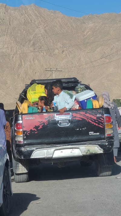 A black Toyota Hilux pickup truck, loaded with personal belongings and several people, is parked on a paved road in Bostan, Pakistan. The vehicle is packed with items including watermelons, a white cooler, and wrapped bundles. At least three children and several adults are visible within the load. The environment is arid and mountainous, with a clear blue sky partially visible in the background. Electrical lines run parallel to the road.