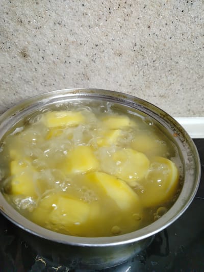 A close-up, overhead shot shows potato chunks boiling in a metal pot. The pot is on a black stovetop, and behind it is a speckled countertop. The potatoes are cut into irregular shapes and are submerged in clear, bubbling water. The water is hot and steamy, indicating the cooking process is well underway. The scene is domestic, likely taking place in a kitchen. There are no people or animals visible in the image. The environment suggests a typical household setting where food preparation is occurring.