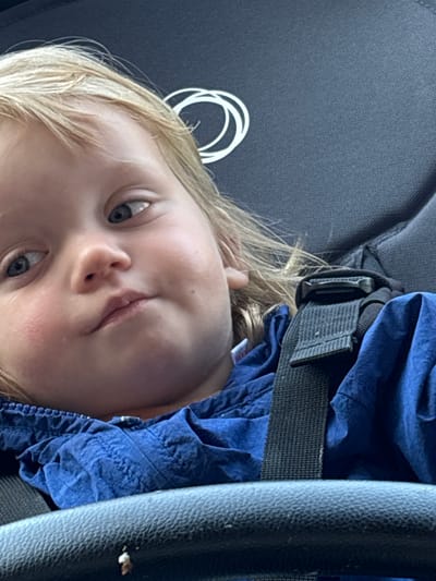 A close-up, tilted, eye-level shot shows a young child with blond hair, lying in a dark grey stroller. The child has fair skin, blue eyes, and a neutral expression. A blue puffy jacket surrounds the child, and black straps are visible, presumably part of the stroller's safety harness. The background includes a dark grey material, possibly the stroller's interior. The location is not discernible from the image provided. Longone al Segrino, Italy is not reflected in this image.