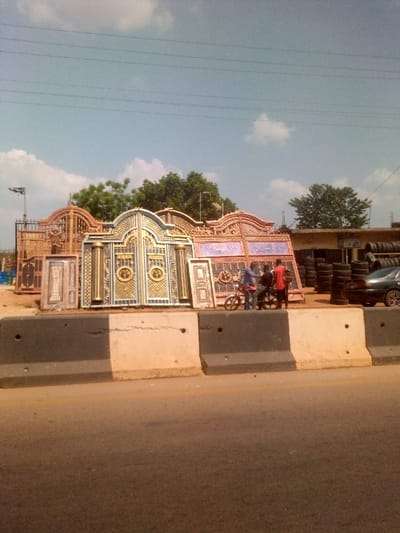 The scene depicts a street-side commercial area in Agbani, Nigeria. In the foreground, a multi-lane asphalt road is separated from the adjacent commercial establishments by a line of concrete jersey barriers, painted alternately black and off-white/beige.
Beyond the barriers, on an unpaved dirt surface, multiple large, ornate metal gates and doors are displayed for sale. These gates exhibit intricate designs, featuring various colors including gold, blue, pink, and black. Some gates have arched tops, while others are rectangular. Notable examples include a prominent blue and gold double gate with circular motifs and a pink/red and gold gate with geometric patterns. Smaller single doors are also visible amongst the larger gates.
To the right of the gate display, three adult males are visible. One individual, wearing a blue long-sleeved shirt and trousers, is standing near a black motorcycle. Another individual, wearing a red short-sleeved shirt and dark trousers, stands to his right, facing the first man. A third individual, wearing a dark shirt and trousers, is positioned between the first two, appearing to engage in conversation. The motorcycle is dark-colored with spoked wheels.
Further to the right, a tire shop operates, with numerous stacks of vehicle tires piled high against a building structure. A dark-colored sedan is partially visible parked near the tire piles.
In the background, mature green trees are present, along with utility poles and power lines spanning across the clear, light blue sky which features scattered white clouds. A street light is also visible on the left side of the image.