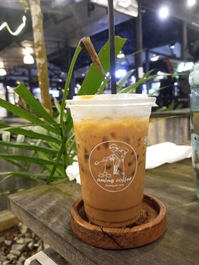 A clear plastic cup containing iced coffee, distinguishable by its light brown color and ice cubes, is centrally positioned on a weathered, light gray wooden table. Condensation is visible on the exterior of the cup. A black plastic straw is inserted, angled slightly left. The cup features a white circular logo depicting a person preparing coffee, with the text "aming coffee Pontianak 1978" underneath. This cup rests on a dark brown, round wooden coaster, which holds a small pool of water from the cup's condensation.
To the immediate right of the cup on the table, a crumpled white napkin is present. A partial view of a light blue or green plastic bottle is visible at the far right edge of the frame. The background is out of focus, depicting a nighttime or dimly lit outdoor setting. Lush green foliage, including broad leaves and a slender tree trunk, occupies the left side and mid-ground. A distinct brown, dried leaf segment points upward behind the coffee cup, adjacent to the straw. Multiple artificial light sources, including hanging fixtures, illuminate a blurred structural background, indicating a commercial establishment. The ground below the table on the left is composed of blurred gravel. The location is an Aming Coffee establishment in Pontianak, Indonesia, as indicated by the cup's branding. No individuals are present in the frame.