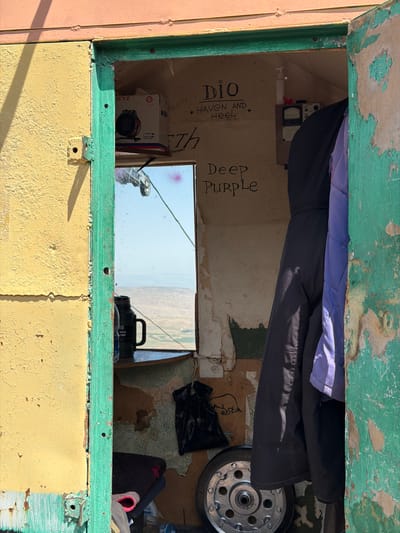The image depicts the entrance to a small, weathered structure with peeling yellow and pink paint on the exterior. The doorway is framed by a bright green, chipped frame. Inside, the walls are unfinished and appear to be made of plywood or similar material, with some water damage and peeling wallpaper.

On the interior wall, there is graffiti. At the top left, a white box with the text "BEYZ" and "3500WATT" is visible. To the right of this, also on the wall, is graffiti that reads "Dio" and "HAVEN AND HEEL" with a small star. Further down, larger graffiti spells out "Deep Purple".

A mirror is positioned on the left side of the interior, reflecting an outdoor scene with a landscape of rolling hills under a clear blue sky. On a shelf below the mirror sits a black thermos.

Two jackets hang in the doorway: one dark navy and one a lighter purple. A black plastic bag is on the floor inside, next to a silver metal wheel with a circular pattern. On the lower right of the wall, a drawing of an animal, possibly a bear, is visible.

The scene appears to be located in an outdoor, possibly remote setting given the landscape visible in the mirror. The overall impression is one of a rustic, utilitarian space, possibly a storage shed or a small shelter, marked by personal touches like the graffiti and hanging clothes. The specific location in Kashka Suu, Kyrgyzstan, suggests a context of a village or rural area, fitting with the observed environment.