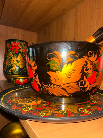 In a dimly lit interior, likely a cupboard or shelf, lie several traditional Russian nesting dolls, known as Matryoshka dolls, and a decorative tray. The subjects are the dolls and tray themselves, painted with vibrant, intricate folk art. The largest Matryoshka doll is visible in the foreground, its black surface adorned with detailed golden foliage and bright red strawberries, characteristic of Khokhloma painting style. To its right, a matching decorative tray shares the same Khokhloma design, featuring floral motifs in red, yellow, green, and orange against a black background. Below the large doll, a smaller Matryoshka doll is peeking out, also decorated with a similar folk art pattern of red berries and yellow flowers. A partially visible golden object, possibly another doll or decorative item, is situated to the far right, catching the light. The scene is static, with no people or obvious activity, suggesting the items are stored or displayed. The lighting is artificial, creating a warm glow and highlighting the glossy finish of the painted items. The setting appears to be a domestic space, given the wooden shelving and the nature of the objects. There is no visible text in the image.