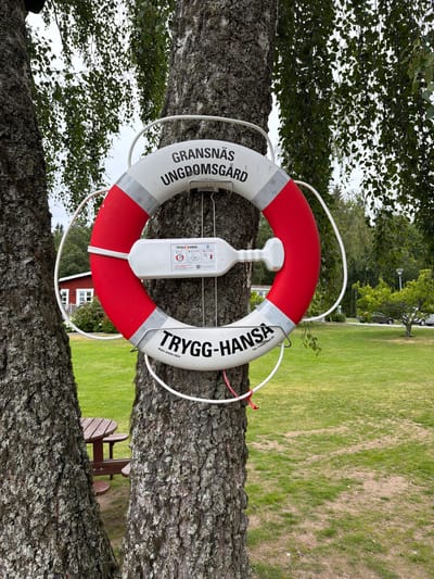 A red and white life preserver is attached to a tree in Aneby, Sweden, near a building and a picnic table. The life preserver has text that reads "GRANSNÄS UNGDOMSGÅRD" and "TRYGG-HANSA". A white plastic box with instructions and diagrams is attached to the life preserver. The background shows a grassy area with trees and a building.