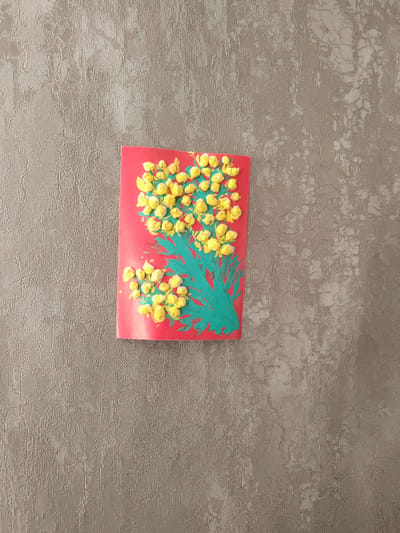 This media file features a handmade craft on a textured, grey wall. The craft is a rectangular piece of red paper with a painted image of a mimosa branch. The mimosa flowers are depicted as clusters of yellow, bumpy spheres, likely made from paint or small craft materials. A green painted stem and leaves extend from the bottom of the red paper, creating a stylized branch. The craft is positioned slightly off-center on the wall, with the majority of the frame occupied by the wall's textured surface, which has a pattern of raised lines and crackle effects. There is no visible text. The lighting suggests an indoor setting, likely during the daytime, given the even illumination without strong shadows. The scene is simple, focusing on the contrast between the vibrant craft and the neutral background. The overall impression is of a child's artwork displayed at home.