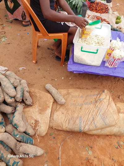 In Awkuzu, Nigeria, a market scene unfolds. In the foreground, a pile of root vegetables, likely yams, lies on a burlap sack. Some of the vegetables have blue markings. To the right, a person, partially visible, is scooping popcorn from a yellow and red bag into a large clear plastic container. Several other bags of popcorn are scattered around the container, which rests on a purple tarp. In the background, a pile of red palm nuts and some green fruits are visible on a stall. The ground is sandy earth, scattered with dry leaves and debris, indicative of an outdoor market environment. A person is seated on an orange plastic chair, their legs and arms visible. Their attire includes a black t-shirt and black leggings, and they wear brown shoes. The overall impression is that of a lively, transactional setting within the Nigerian community.