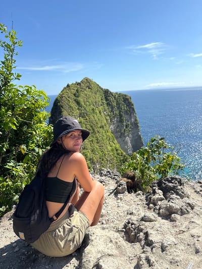 A woman sits on a rocky outcrop near a cliff in Bunga Mekar, Indonesia. The woman wears a black tank top, olive-green shorts, and a black bucket hat. A black bag rests beside her. Behind her is a steep, green-covered cliff and the blue ocean. The sky is clear and blue. There are small monkeys resting on the rock, next to the woman. Green foliage is visible in the foreground.