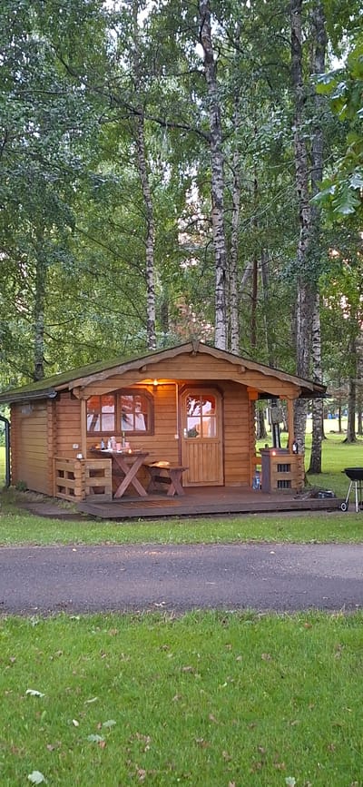 The image depicts a charming log cabin nestled amidst a lush, green forest. The cabin has a rustic wooden exterior with a pitched roof and a covered porch. In front of the cabin, a wooden picnic table and benches are set up, suggesting an outdoor dining area. A barbecue grill is visible to the right of the cabin. The surrounding environment is characterized by dense trees, with sunlight filtering through the leaves, creating dappled light. The ground is covered in grass, and a paved path runs in front of the scene. The overall atmosphere is serene and idyllic, evoking a sense of peace and tranquility.
