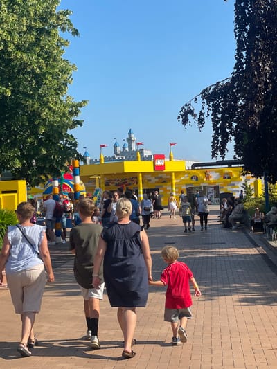 The image depicts a sunny day in Billund, Denmark, at the LEGO House or a similar LEGO-themed attraction. The foreground features a family walking away from the camera along a brick pathway. A woman in a light blue top and light-colored shorts is on the left, followed by a person in a dark green t-shirt and white shorts, and a woman in a dark blue dress. A young boy in a red t-shirt and grey shorts is walking hand-in-hand with the woman in the dark blue dress.

In the mid-ground, a brightly colored LEGO structure stands tall, adorned with red, blue, and yellow bricks. Behind it, a yellow building with a prominent red "LEGO" logo is visible, resembling a storefront or attraction entrance. This building is constructed with LEGO bricks and features a castle-like facade with blue turrets and red flags.

The background shows a clear blue sky and more structures resembling LEGO castles, with more turrets and flags. Several people are walking around the attraction, browsing the shops, or sitting on benches. Trees with green and dark foliage frame the scene on either side. The overall atmosphere appears to be one of a lively, family-friendly outdoor venue.
