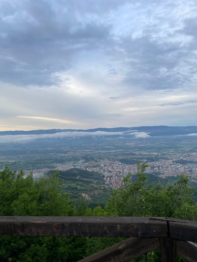A high-angle, full shot captures a view of Delchevo, Bulgaria, from a lookout point. The foreground is dominated by a dark, weathered wooden railing. Beyond the railing, lush green trees form a dense canopy, their leaves slightly blurred. In the middle ground, the city of Delchevo sprawls across a valley, its buildings characterized by red-tiled roofs. The city is nestled among rolling green hills. In the background, a mountain range stretches across the horizon, partially obscured by a band of soft, white clouds that hug the lower slopes. The sky above is overcast with layers of gray and white clouds, with a faint strip of lighter sky visible near the horizon.