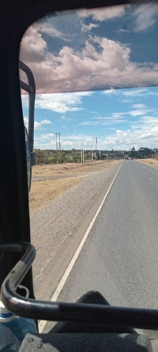 A view from inside a vehicle shows a long, straight road stretching into the distance under a blue sky with scattered clouds. The road is asphalt with a white dividing line. Vegetation lines the sides of the road, including grass and some trees with power poles. Vehicle interior elements such as the windshield frame and metal bars are visible, partially obscuring the view of the sky and road.