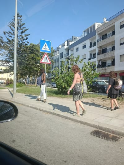 The image depicts a street scene in Sines, Portugal, under a clear blue sky. In the foreground, a side mirror of a vehicle is visible on the left. The street itself is paved, with a sidewalk running alongside it.

On the sidewalk, three individuals are walking. The closest to the viewer is a person wearing a light-colored shirt, dark shorts, and dark shoes, carrying a dark backpack. To their left and slightly behind is another person, a woman, wearing a light-colored top, camouflage shorts, and light-colored slip-on shoes, carrying a patterned bag over her shoulder. Further to the left and ahead of the first person is another individual, a man, wearing a hat, sunglasses, white pants, and brown shoes, with a dark backpack slung over his shoulder.

Two traffic signs are visible on the sidewalk. The upper sign is a blue square with a white pedestrian symbol, indicating a pedestrian crossing. Below it is a red-bordered triangular sign with two black arrows pointing upwards, indicating a narrowing of the road.

Behind the sidewalk is a grassy area with trees and bushes. In the background, a modern multi-story white building with balconies and dark-framed windows is prominent. Another building, partially visible to the left of the white building, appears to be older with yellow and beige facades. Several parked cars are visible on the street in front of the buildings, including a black SUV. A dog is also visible near the people on the sidewalk.