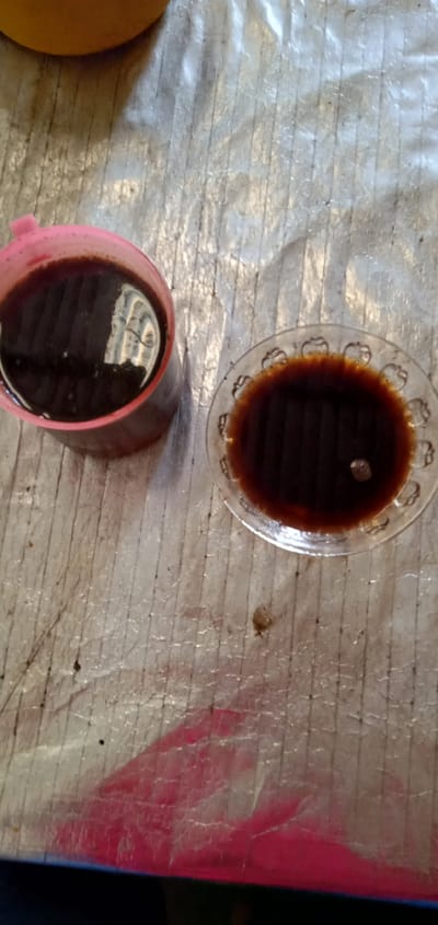 The image shows two containers, a pink mug and a small glass bowl, both filled with a dark liquid, likely coffee or tea, resting on a textured, slightly stained surface. The background is indistinct, suggesting an indoor setting. The picture was taken in Sumput, Indonesia.