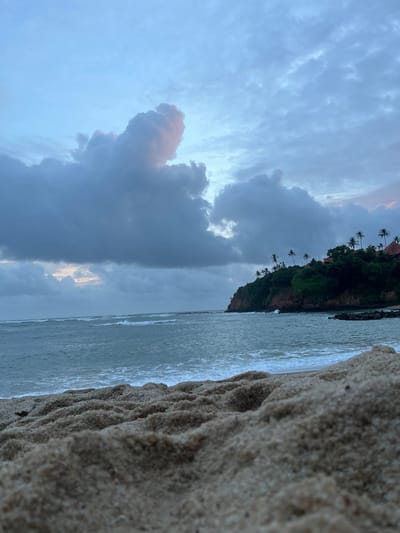 This is a landscape photograph taken at Batawala, Sri Lanka. The image depicts a serene beach scene with the ocean on the left, a cliffside covered in lush green vegetation and palm trees in the middle, and a dramatic sky filled with clouds on the right. The foreground is dominated by coarse sand, with wave patterns visible along the edge where the sand meets the water. The ocean appears calm, with gentle waves breaking on the shore.

The cliffside is a prominent feature, with a dense covering of trees and palm trees scattered across its slopes, suggesting a tropical location. A few buildings with reddish-brown roofs are partially visible amongst the trees, hinting at human habitation or a resort.

The sky is filled with a variety of clouds, ranging from dark, brooding storm clouds to lighter, wispy formations. The presence of a bright patch of light peeking through the clouds suggests either the sun setting or rising, or the clouds parting to reveal the sky beyond. The overall mood of the image is tranquil and slightly melancholic, perhaps due to the overcast sky and the approaching twilight. The time of day appears to be either early morning or late evening.

There is no visible text in the image. The composition, with the sand in the foreground leading the eye towards the ocean and the cliffside, creates a sense of depth and natural beauty.