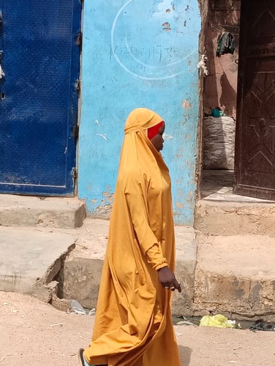 The image depicts an outdoor street scene in Zango, Nigeria, under bright daylight. A single individual, appearing to be a dark-skinned female, walks in profile from right to left across the frame. She wears a full-length, loose-fitting mustard yellow or deep ochre abaya/jilbab, with a bright red under-scarf visible beneath the yellow head covering. Her visible right hand, with dark skin, displays two rings on the ring finger: one gold-toned and one silver-toned. Her left foot, partially visible, wears a dark-colored sandal or flip-flop.

The background consists of building exteriors. To the left is a partially visible dark blue metal door with a latch. Adjacent to this door is a weathered, light blue painted wall exhibiting peeling paint and minor damage. Faint white handwritten text, including numbers such as "070204143," is inscribed within a partial circular outline on this blue wall. To the right of the blue wall, a weathered, reddish-brown or mud-colored wall is present. This wall contains a partially open dark brown wooden door, revealing a darker interior. A dark green or brown item, possibly a bag, hangs from the brown wall above the doorway, with a white plastic object beneath it. Multiple uneven, chipped concrete or stone steps lead up to both the blue wall's base and the brown doorway. The ground beneath the subject's feet is dry, light brown dirt or earth, scattered with small pebbles and pieces of refuse, including a crumpled yellow plastic bag and white plastic fragments.